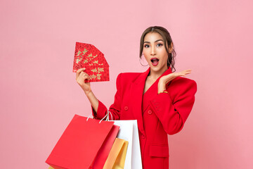 Surprised beautiful Asian woman in red jacket carrying shopping bags and holding Chinese new year envelopes or Ang Pao in pink background, foreign text translation as every thing comes true