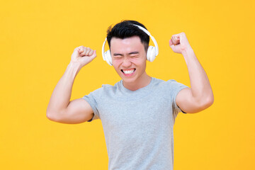 Happy Asian man wearing headphones listening and enjoying music with arms raised on yellow background studio shot