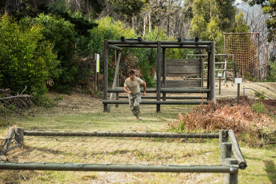 Male trainee sprinting through wooden hurdles and climbing rope net over mud patch on forest course - Powered by Adobe