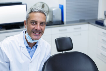 Male dentist leaning on black dental chair in clinic room with monitor, oscillating fan, copy space