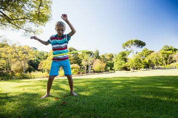 Boy wearing striped polo shirt standing barefoot on grass spinning hula hoop at park, copy space