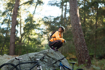 Female cyclist crouching on mossy rock in pine forest adjusting bike helmet beside mountain bikes