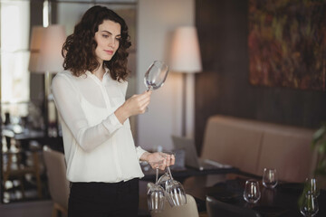 Female server wearing white blouse examining wine glasses under table lamp light in restaurant