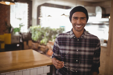 Obraz premium Man holding eyeglasses leaning against wooden counter wearing beanie and plaid in cafe, copy space