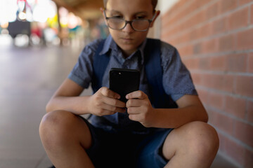School-age boy sitting on walkway near red brick wall using smartphone with eyeglasses and backpack