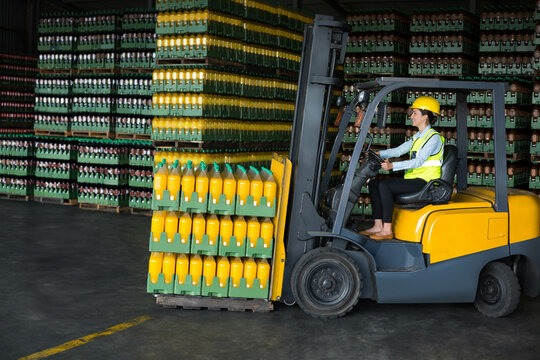 Forklift operator wearing helmet, hi-vis vest is lifting pallet of orange juice crates in warehouse - Powered by Adobe