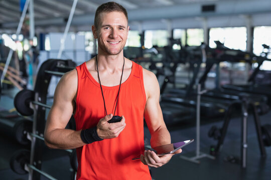 Fototapeta Mid adult trainer wearing athletic top holding stopwatch tablet in gym near weight benches, mirrors