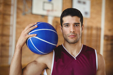 Male basketball player in jersey holding blue basketball on shoulder inside gym under mounted hoop