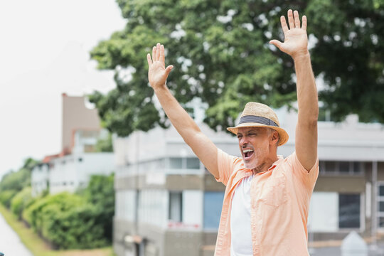 Senior man raising arms shouting beside canal path wearing straw fedora hat peach shirt, copy space