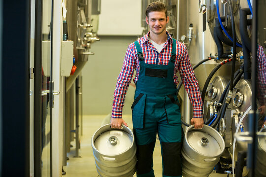 Man carrying two beer kegs walking down brewery corridor flanked by stainless steel tanks
