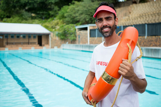 Male lifeguard with red visor holding orange rescue buoy with rope beside pool lanes, copy space - Powered by Adobe