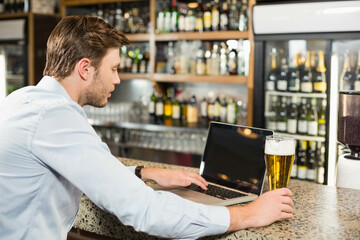 Man is working on laptop at stone bar counter with pint of beer near liquor shelves