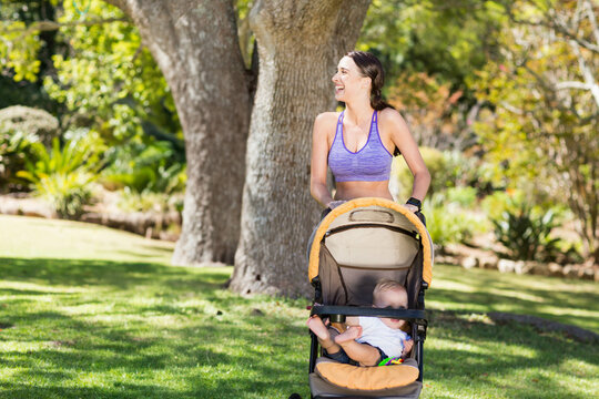 Fototapeta Mother and infant in sports attire pushing stroller across park under sunlight with fitness tracker