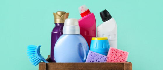 Box with cleaning supplies and bottles of detergent on turquoise background, closeup