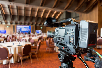 A professional video camera on a tripod set up in a spacious conference hall, facing round tables...