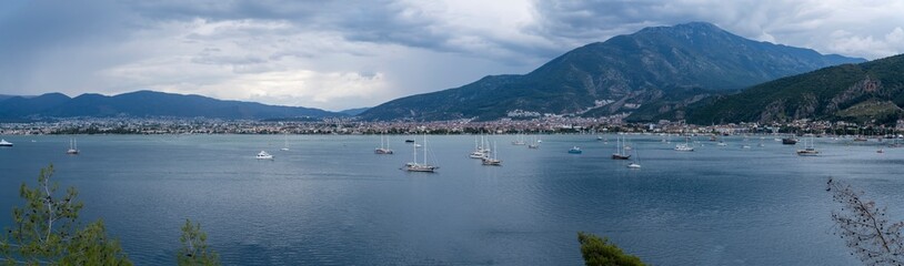 Panoramic view of Fethiye town lively harbor, lined with boats and yachts, set against a scenic coastal townscape and calm Mediterranean waters in daylight.