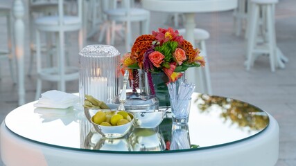An elegant table setting with a floral centerpiece, olives, pickles, and drink stirrers on a reflective surface, with blurred chairs and tables in background