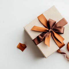 A beige square gift box adorned with a brown and burnt orange satin ribbon, tied in a classic bow, sits on a white background; small ribbon scraps are nearby