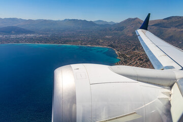 Airplane wing and engine in view as the plane descends over a coastal town with turquoise waters and surrounding mountains.
