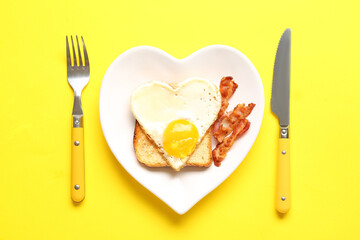 Plate with tasty bacon, toast, heart made of fried egg and cutlery on yellow background