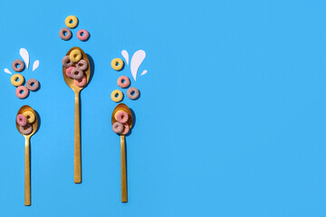 Spoons with colorful sweet cereal rings and paper milk drops on blue background