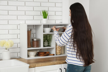 Pretty young woman putting dishes onto shelves in light kitchen, back view
