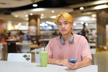 Thai gay teen man with headphones using phone and drinking iced latte green tea in modern cafe
