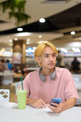 Thai gay teen man with headphones using phone and drinking iced latte green tea in modern cafe
