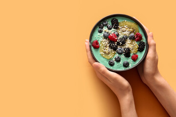 Female hands and bowl of tasty spirulina smoothie with berries on orange background