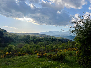 Scenic view of rolling hills, mountains, and a cloudy sky under sunlight. Mountain range with wildflowers. Landscape with Rolling Hills and Cloudy 
