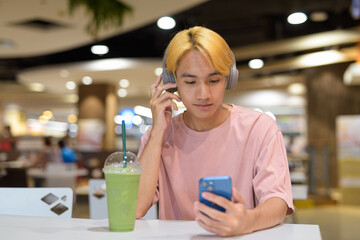 Thai gay teen man with headphones using phone and drinking iced latte green tea in modern cafe