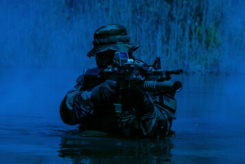 Soldier in camouflage gear wading in water, holding a rifle with tactical equipment, surrounded by dense vegetation under a blue-tinted light.