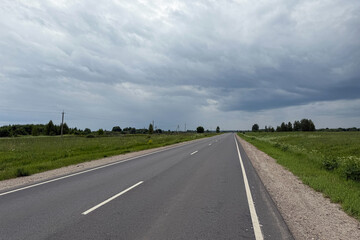 Asphalt road in the countryside running through green fields on a cloudy day. Rural landscape with road and storm clouds