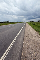 Asphalt road in the countryside running through green fields on a cloudy day. Rural landscape with road and storm clouds