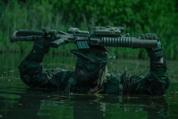 Camouflaged soldier with face paint held rifle above head while wading through water in a dense forest environment under daylight.