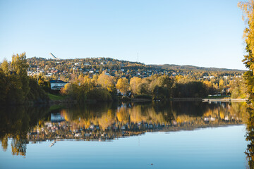 Fototapeta premium Autumn reflections dance on the still waters of Holmendammen Lake in Oslo, surrounded by colorful trees and hillside homes under a crisp, cloudless sky