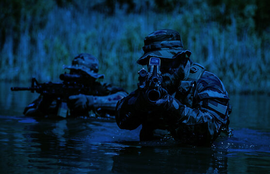 Two armed soldiers in camouflage uniforms navigate through water with rifles aimed forward, under a low-light environment and surrounded by vegetation.