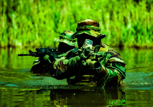 Soldiers in camouflage uniforms with rifles navigate through water in a dense green vegetation setting, demonstrating stealth and tactical movement.