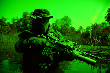 Soldier in camouflage with a rifle crouches in water surrounded by dense vegetation, wearing a hat and gloves, during low-light conditions.