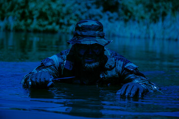 Man in camouflage clothing and hat crouching in water holding a knife with foliage in the background.