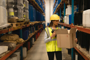 Female clerk wearing safety vest examining box while scanning barcodes between metal shelves