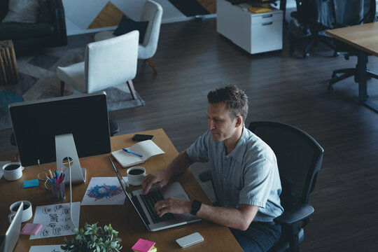 Mature male office worker typing on laptop with monitor and coffee cups in office, copy space - Powered by Adobe