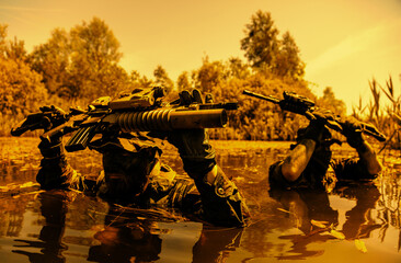 Two camouflaged soldiers wade through water holding rifles above their heads, with trees in the background on a clear day.