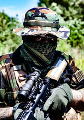 Soldier with camo gear and face covering, holding a rifle with optics in a green, wooded outdoor setting under clear blue sky.