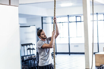 Man gripping and climbing thick rope in cross-training gym with squat rack and plyometric box
