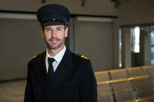 Mature male pilot standing near seats in airport lounge wearing peaked cap with striped epaulets