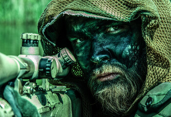 A sniper in a camouflage netting hood with face paint peers through a rifle scope in a dense green environment.