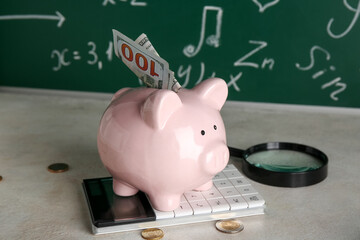 Piggy bank with money, calculator and magnifier on table against blackboard. Student loan concept