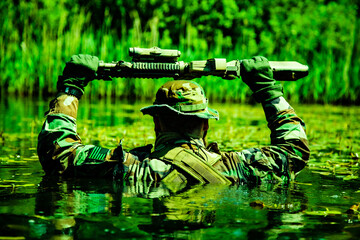 Soldier in camouflage holding a rifle above water while wading through a green swampy area surrounded by vegetation.