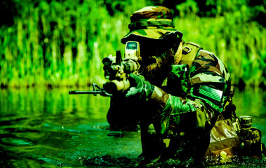 Soldier in camouflage uniform and hat aiming a rifle with a scope while wading through a green...